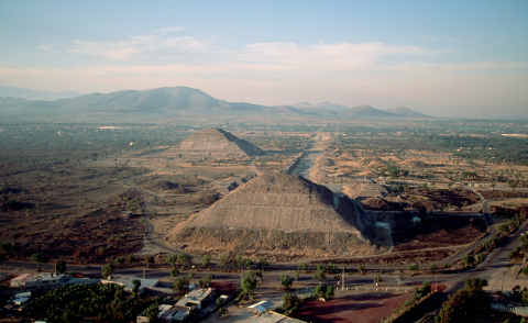Aerial view of the Teotihuacan pyramids, the Pyramid of the Sun and the Pyramid of the Moon, connected by the Avenue of the Dead, under a blue sky with distant mountains.