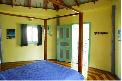 A bedroom in the "Bocas del Toro Getaway" features yellow walls, a four-poster bed with blue bedding, and a window with dark curtains. A colorful door is visible, leading to another room.