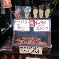 En gadesælgers udstilling i Japan præsenterer forskellige snacks, herunder tørret blæksprutte, pakkede godbidder og kegleformede slik, alt sammen prissat med håndskrevne skilte.
