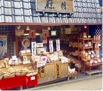 En charmerende japansk butik med tegltag præsenterer et udvalg af snacks og lækkerier på hylder og en trædisk, hvilket skaber en indbydende og traditionel butiksfacade.