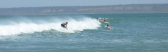 Cabo Ledo Surfing Weekend A surfer rides a wave while two others float nearby, with a forested landmass visible in the distance.