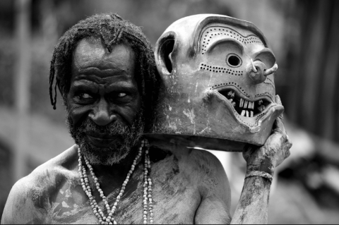 A striking black and white portrait captures a man with dreadlocks and a beaded necklace holding a carved, mask-like face beside his head.