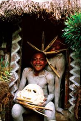 Certainly! Here is a description of the image based on the provided details and what is visible:A smiling person painted white holds a mask and sticks in front of a hut with patterned decorations, while another person with a spiky headpiece is partially visible behind them. 