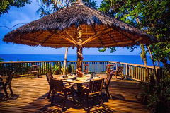 A patio with a large thatched umbrella on a wooden deck overlooks the ocean, featuring a dining table and chairs for an inviting outdoor meal.