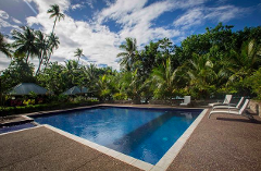 A picturesque scene of a tropical swimming pool surrounded by lush greenery, palm trees, and lounge chairs under a partly cloudy blue sky.