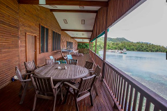 A spacious wooden porch with a round table and chairs overlooks a scenic ocean view with a boat and lush green islands in the distance.