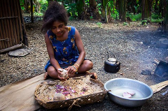 A woman in Papua New Guinea prepares food outdoors, peeling potatoes into a woven basket, with a kettle and a bowl of water nearby.