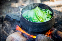 A black pot filled with food wrapped in green leaves sits on a fire, with smoke rising, suggesting cooking in a traditional setting.