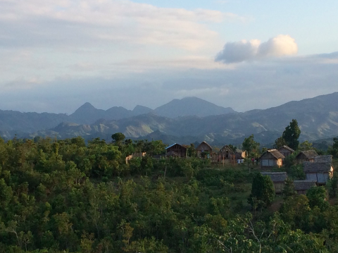 A scenic view of a hillside village with traditional houses, set against a backdrop of layered mountains and a partly cloudy sky.