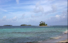 The image shows a tropical beach with clear turquoise water and three small islands in the distance under a cloudy sky.