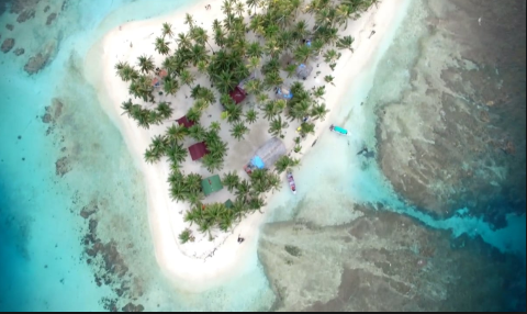 An aerial view of a tropical island with white sand beaches, palm trees, small buildings, and boats in crystal-clear turquoise waters.