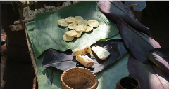 Food arranged on banana leaves: small, round flatbreads on a green leaf, and chunks of grilled food on a dark purple leaf, with a woven basket and a small cup nearby.