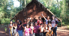 A group of people, including adults and children, stand in front of a mud hut, some waving, with a woman holding a guitar, possibly a cultural exchange or visit.
