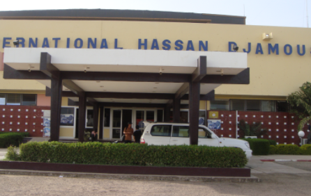 This is a low-angle shot of the entrance to the International Hassan Djamous Airport in N'Djamena, Chad. The entrance features a modern canopy and a white car parked in front of lush greenery.