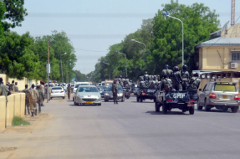 A column of soldiers in the back of pickup trucks branded "GMIP" travels down a city street lined with vehicles and pedestrians.