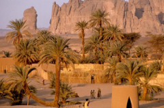 This image shows a desert oasis with palm trees, buildings, and people, set against a backdrop of rocky cliffs and desert landscape.