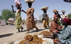 A bustling street scene in Africa featuring women carrying items on their heads, and others sitting and selling goods on the ground.