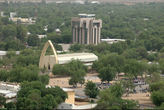 An aerial shot captures a cityscape with lush greenery, featuring a prominent building with a large arch and a taller, modern structure in the background.