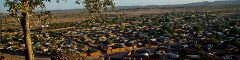 A sprawling village with a mix of traditional dwellings and brick structures spreads across a dry, expansive landscape, viewed from an elevated vantage point.