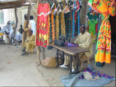 A tailor works on a sewing machine at an outdoor shop with colorful dresses on display, as other people watch in the background.