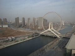 Here's a one-sentence description of the image:A view of the Tianjin Eye Ferris wheel in Tianjin, China, spanning the Hai River, with city buildings in the background, on a slightly hazy day.