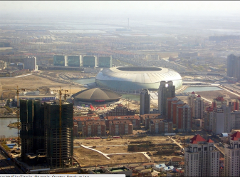 An aerial view shows the Tianjin Olympic Center Stadium surrounded by urban development, including buildings under construction and residential areas.