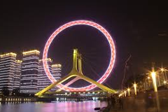 A brightly lit Ferris wheel stands prominently over a river at night, with illuminated buildings in the background, creating a vibrant urban scene.
