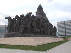 A large bronze monument depicts a group of people and horses, atop a light-colored stone base with buildings in the background.