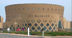 A large, cylindrical, brown building with diamond-shaped windows and Chinese characters on the facade stands next to a street with traffic signals and decorative sculptures.