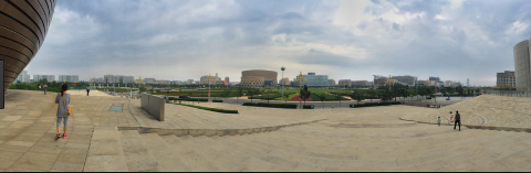 A wide shot of a cityscape with a woman in the foreground, stone steps leading down to a park with buildings in the background, and a cloudy sky above.
