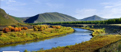 A scenic view of a river meandering through a grassy landscape with hills and mountains in the background under a blue sky.