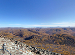 A scenic view of rolling hills covered in autumn foliage under a clear blue sky, captured from a stone overlook with a chain-link fence.