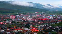 A panoramic view of a town nestled in a valley, featuring red-roofed buildings, green hills, and a misty mountain range in the background.