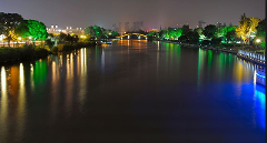 Huzurlu bir gece manzarası, kıyılarında yemyeşil bitki örtüsünün bulunduğu, aydınlatılmış şehir manzarasını ve köprüyü yansıtan bir nehri sergiliyor.