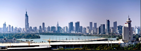 A panoramic view of Nanjing, China shows a skyline of modern skyscrapers across a lake and a low stadium in the foreground.