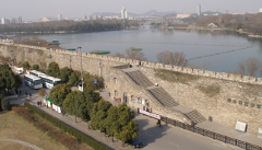 An elevated view captures a stone wall with staircases, flanked by trees and buses, alongside a calm lake with distant city buildings.