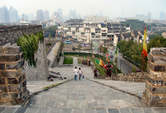 From atop a stone stairway, a couple descends towards a bustling street in a city with a blend of historic and modern architecture, decorated with flags and statues.
