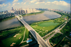 This aerial shot showcases a long, modern bridge over a river, with a city skyline in the background and lush green golf courses and a lake on either side.