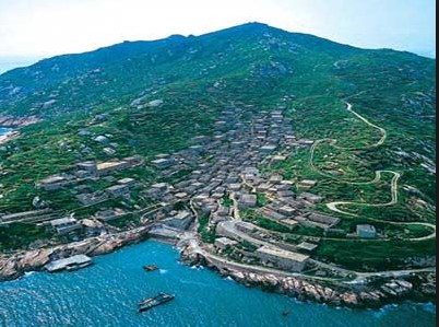An aerial view shows a deserted village nestled on a lush island with a mountain backdrop, bordered by a rocky coastline and turquoise sea.