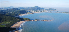 An aerial view shows a scenic coastline with sandy beaches, lush green hills, and calm blue water, under a clear sky.