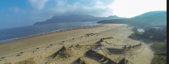 Panoramic view of a crowded sandy beach with mountains in the background and elaborate sand structures in the foreground.