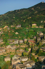 A village covered in green vegetation clings to the slopes of a hillside, with a structure visible at the top of the hill.