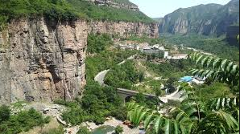 Here's a description of the image:A picturesque view of a town nestled in a valley surrounded by towering cliffs and lush green vegetation, featuring a winding road and an arched bridge.