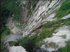 Here's a description of the image:A high-angle shot shows a steep, rocky mountainside with sparse green vegetation clinging to its surface, leading down to a rocky valley floor.