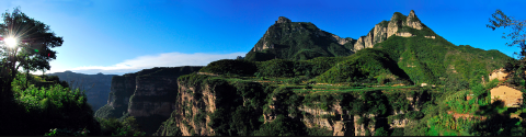 A scenic panoramic view shows a lush green mountain range under a bright blue sky, with sunlight shining through a tree on the left.
