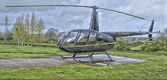 A sleek black helicopter rests on a concrete pad amidst a lush green landscape under a cloudy sky.