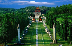 A long path lined with statues and pillars leads to a traditional Chinese building amidst lush greenery and a clear blue sky.