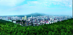 A wide-angle shot shows a cityscape nestled against a backdrop of rolling mountains and lush greenery.