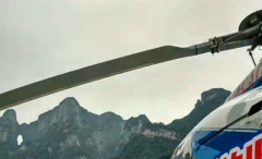 A helicopter blade is silhouetted against a backdrop of rugged mountains under a cloudy sky.