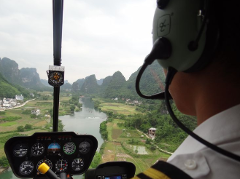A view from a helicopter cockpit, showcasing the pilot in headphones and the instrument panel, with a breathtaking panorama of karst mountains, a winding river, and rural landscapes stretching into the distance.
